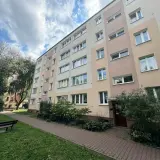 Blok mieszkalny z oknami i balkonami, z zielenią i ławką przed budynkiem.