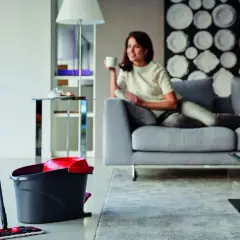 Mop i wiadro stoją na podłodze w salonie, gdzie kobieta odpoczywa na kanapie.