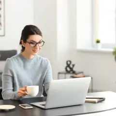 Kobieta w okularach pije kawę, pracując na laptopie przy biurku.