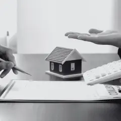 Dwie osoby przy stole, jedna pisze, druga trzyma kalkulator, obok model domu i dokumenty.