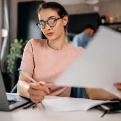 Kobieta w okularach pracuje przy laptopie i dokumentach.