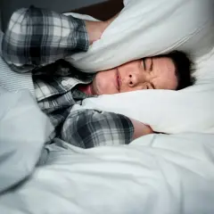 Kobieta w piżamie zakrywa uszy poduszką, leżąc w łóżku.