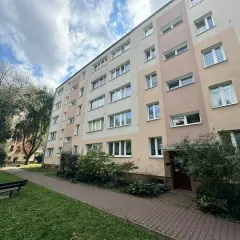 Blok mieszkalny z oknami i balkonami, z zielenią i ławką przed budynkiem.