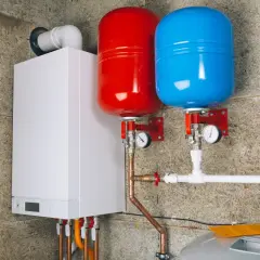 Biały piecyk gazowy z dwoma zbiornikami (czerwonym i niebieskim) oraz instalacją hydrauliczną.