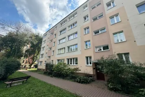 Blok mieszkalny z oknami i balkonami, z zielenią i ławką przed budynkiem.