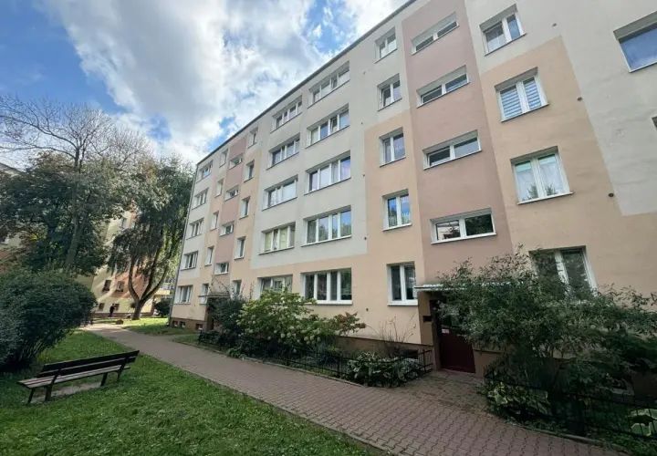 Blok mieszkalny z oknami i balkonami, z zielenią i ławką przed budynkiem.