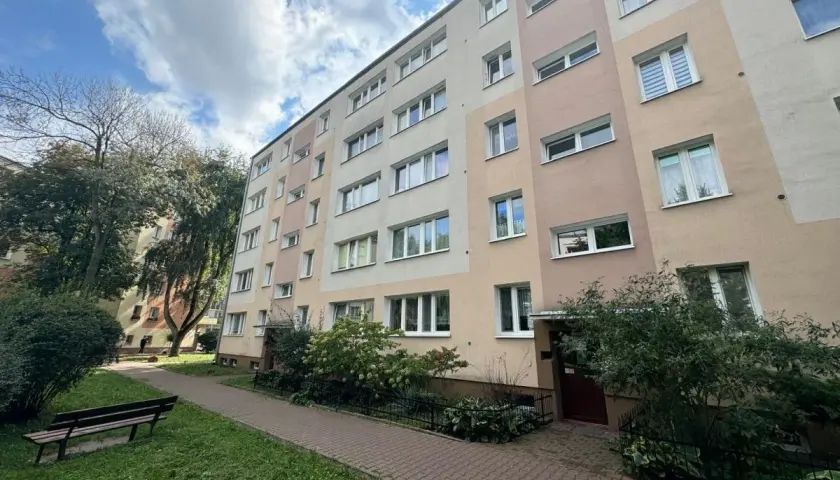Blok mieszkalny z oknami i balkonami, z zielenią i ławką przed budynkiem.