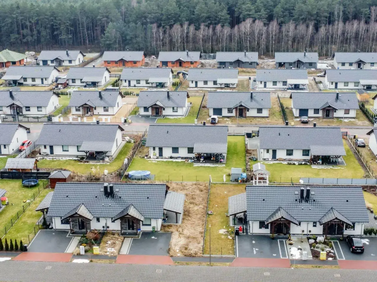 Widok z lotu ptaka na osiedle nowoczesnych domów jednorodzinnych z szarymi dachami, otoczonych lasem.