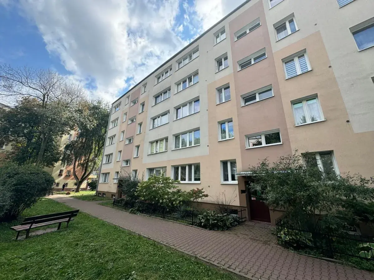 Blok mieszkalny z oknami i balkonami, z zielenią i ławką przed budynkiem.