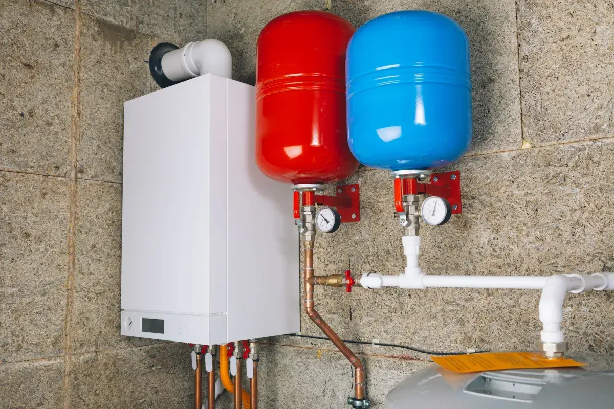 Biały piecyk gazowy z dwoma zbiornikami (czerwonym i niebieskim) oraz instalacją hydrauliczną.